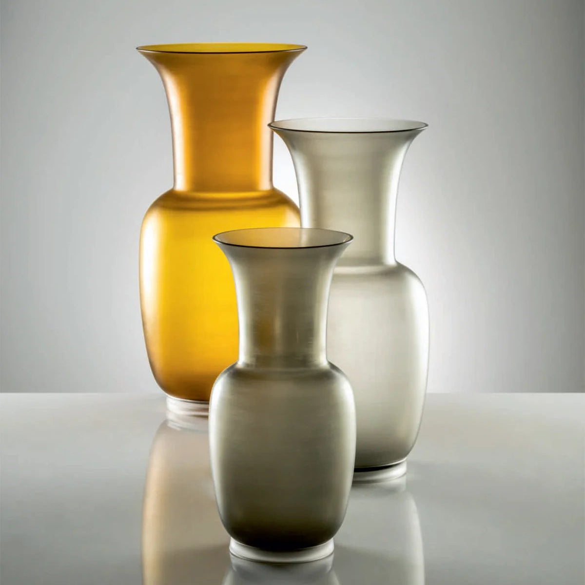 Three hand-blown glass vases of different sizes and colors arranged on a reflective surface against a light gray background. The vase in the back left is translucent amber-yellow, while the two vases in the front and middle are translucent smoky gray.