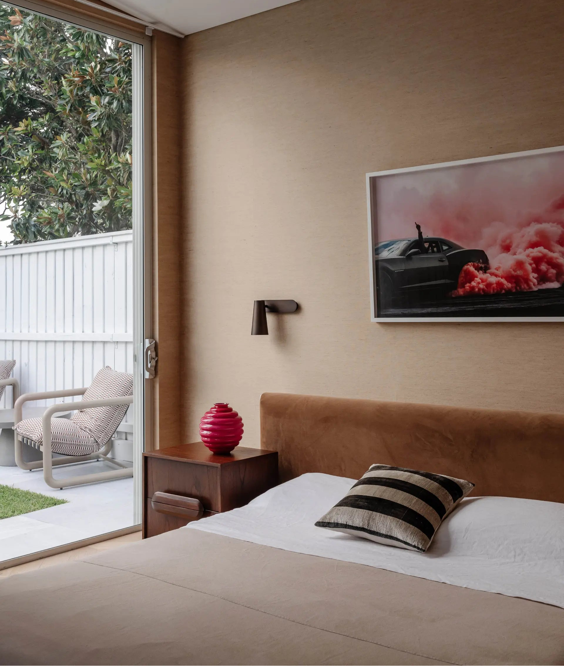 A modern bedroom interior featuring a bed with beige bedding, a brown headboard, a striped accent pillow, a dark wood nightstand with a red decorative object, and a framed wall art print depicting a black sports car against a pink and red background.